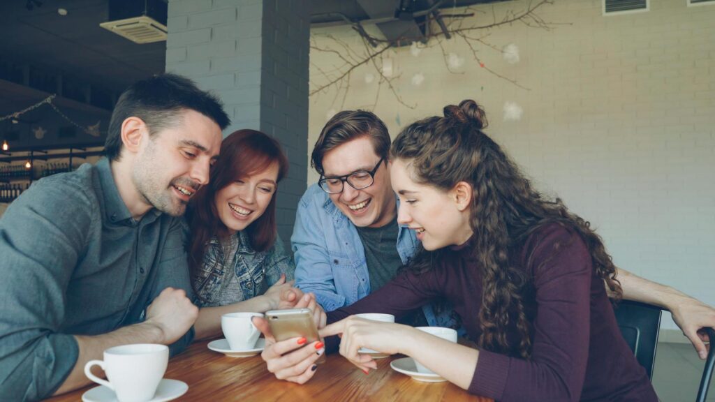 Privathaftpflicht junge erwachsene – Erste eigene Versicherung (Symbolbild: Vier Freunde lachen und tauschen sich bei einer Tasse Kaffee in einem Café aus.)